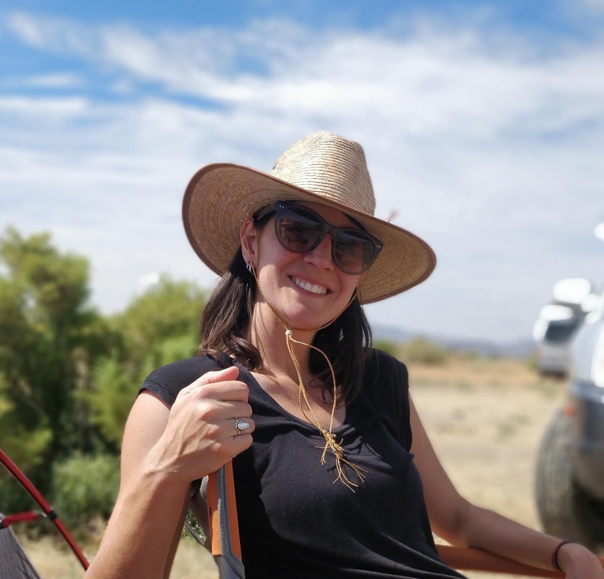 Smiling person outside in desert, wearing black sleeveless top, sunglasses, and a tan sunhat.